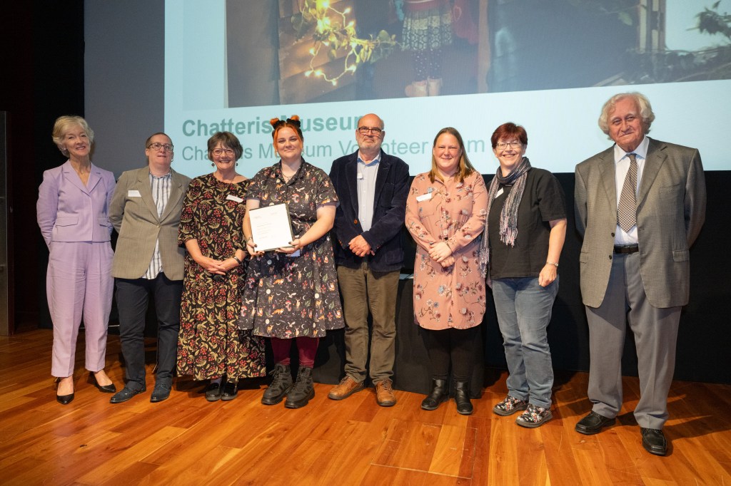 A photo of the 6 Chatteris Museum volunteers standing on the stage where the presentation was held, just after receiving the award. Mr Marsh joining us on the right and Sarah Saunders to the left.