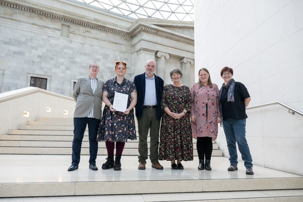 A photo of 6 of the museums volunteers standing on the internal steps of the British Museum holding our certificate with pride. 